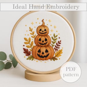 Peut inclure: Une broderie à la main représentant trois citrouilles empilées avec des visages souriants, entourées de feuilles d'automne et d'étoiles. La broderie est placée dans un cercle en bois, avec le texte "Ideal Hand Embroidery" en haut.