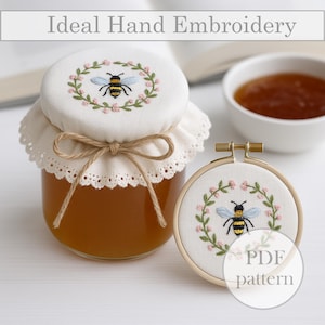 May include: A glass jar of honey with a white fabric lid, hand-embroidered with a bee and floral wreath design. The lid is tied with twine. A small embroidery hoop with a matching bee design is next to the jar. The text "Ideal Hand Embroidery" is at the top.