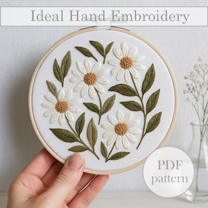 May include: Hand-embroidered daisy design in a wooden hoop. The white daisies with brown centers and green leaves are stitched on white fabric. The text "Ideal Hand Embroidery" is at the top. The text "PDF pattern" is at the bottom.