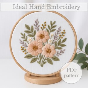 May include: A hand-embroidered floral design in a wooden hoop. The embroidery features three peach-colored flowers with detailed centers, surrounded by green leaves and small white flowers. The text "Ideal Hand Embroidery" is at the top.