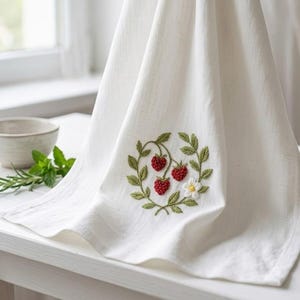 May include: White kitchen towel with an embroidered design of three red strawberries, green leaves, and a small white flower. The towel is folded and draped, showcasing the detailed embroidery. The towel is made of a soft, textured fabric.