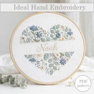 Può includere: Un telaio da ricamo rotondo con un motivo floreale a forma di cuore. Il design include fiori e foglie ricamati blu, bianchi e verdi, con il nome "Noah" cucito al centro. Il testo "Ideal Hand Embroidery" è in alto.