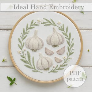 May include: Hand-embroidered garlic design on white fabric, framed in a wooden hoop. The artwork features three garlic bulbs and cloves, surrounded by green leaves and small white flowers. The text "Ideal Hand Embroidery" is at the top, and "PDF pattern" is at the bottom.