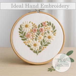 Puede incluir: Bordado a mano con diseño floral en forma de corazón en un aro de madera. El diseño incluye margaritas, rosas y hojas en blanco, rosa y verde. El texto "Ideal Hand Embroidery" está en la parte superior.