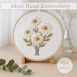 Op de afbeelding: Een handgeborduurd madeliefjesboeket in een houten borduurring. De witte madeliefjes met gele centra en groene bladeren worden getoond op een witte stof. De tekst "Ideal Hand Embroidery" staat bovenaan. De borduurring staat op een houten standaard.