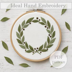 Puede incluir: Un bastidor de bordado con tela blanca con un diseño de corona de hojas y bayas verdes. El texto "Ideal Hand Embroidery" está en la parte superior y una etiqueta "PDF pattern" en la parte inferior derecha. Hojas verdes dispersas alrededor del bastidor.