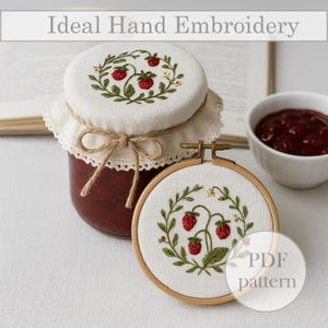 Pode incluir: Um frasco de geleia com uma tampa de tecido branco, amarrada com barbante, e um desenho de morango bordado. Um bastidor de bordado de madeira com um desenho de morango correspondente está ao lado do frasco. O texto "Ideal Hand Embroidery" está no topo.