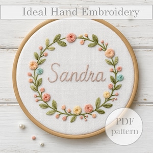 May include: Embroidered hoop art with a floral wreath and the name "Sandra" in the center. The hoop is made of wood and has a white fabric background. The flowers are in shades of pink, yellow, and blue.