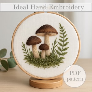 Könnte beinhalten: Handgesticktes Kunstwerk mit drei braunen Pilzen und grünen Blättern auf cremefarbenem Stoff, eingefasst in einen hölzernen Stickrahmen. Der Text "Ideal Hand Embroidery" steht oben. Ein Holzständer stützt den Rahmen. Die Worte "PDF pattern" stehen in einem Kreis.