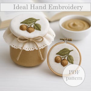 Puede incluir: Un tarro de cristal con tapa color crema, adornado con un diseño bordado de hojas verdes y nueces marrones, atado con hilo. Un pequeño bastidor de bordado con un diseño similar está cerca. El texto "Ideal Hand Embroidery" está en la parte superior.