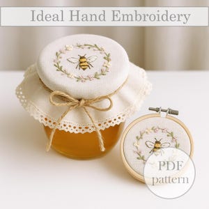 May include: A honey jar with a fabric lid, hand-embroidered with a bee and floral wreath. The jar is tied with twine and a lace-edged fabric. An embroidery hoop with a similar bee design is also visible. The text "Ideal Hand Embroidery" is at the top.