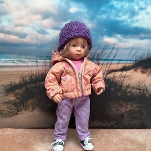 May include: A doll wearing a pink floral print puffer jacket with a purple knitted hat, purple corduroy trousers, and white trainers. The doll is standing against a sandy beach background.