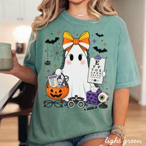 May include: A sage green t-shirt featuring a graphic of a white ghost wearing a candy corn bow. The ghost holds an eye chart with letters E, F, P, T, O, Z, L, P, E, D, P, E, C, F, D, E, D, F, C, Z, P and a pumpkin bucket filled with eye care items.