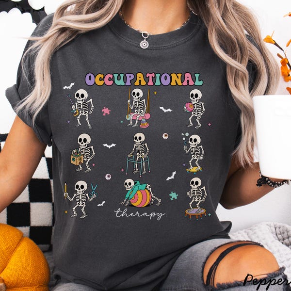 Può includere: T-shirt grigio scuro con un design colorato di figure di scheletri impegnate in attività di terapia occupazionale. La parola "OCCUPATIONAL" è arcuata sopra gli scheletri, con "therapy" scritta sotto. Pipistrelli e altre piccole forme sono sparse intorno al design.