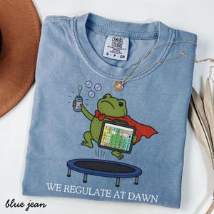 Puede incluir: Una camiseta azul con el gráfico de una rana verde con capa roja, saltando en un pequeño trampolín mientras sostiene una varita de burbujas y un tablero de comunicación. El texto "WE REGULATE AT DAWN" aparece impreso en letras blancas debajo.