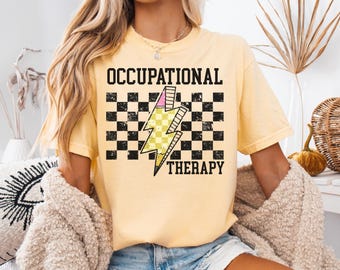 May include: Butter yellow t-shirt with the words "OCCUPATIONAL THERAPY" in black block letters. A yellow and pink lightning bolt graphic is centered on a black and white checkered background. The shirt is a casual style.
