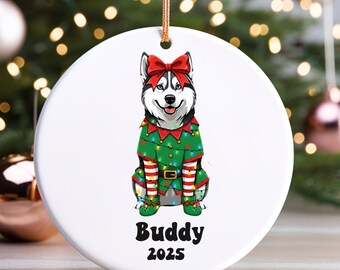 May include: A white ceramic Christmas ornament featuring a Siberian Husky wearing an elf costume and a red bow. The ornament has the text "Buddy 2025" in black. The dog is wearing a green elf suit with red and white striped stockings.