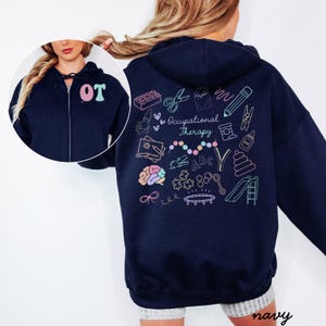 May include: Navy blue zip-up hoodie with the letters "OT" in pink and teal on the chest. The back features colorful line art illustrations related to occupational therapy, including scissors, a brain, and a slide. The word "navy" is at the bottom.