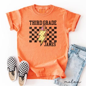 Op de afbeelding: Een oranje t-shirt met de woorden "THIRD GRADE JAMES" in zwarte blokletters. Een gele en roze bliksemflitsgrafiek staat in het midden, tegen een zwart-witte geruite achtergrond. Zwart-wit geruite instappers staan onderaan.