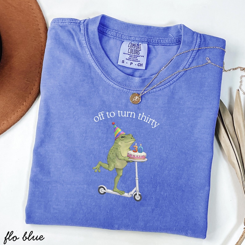 May include: A blue Comfort Colors t-shirt with the text "off to turn thirty" and a graphic of a frog riding a scooter with a birthday cake. The shirt also has a small tag and a gold necklace.