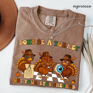 May include: Espresso-colored t-shirt with the text "DONT BE A TURKEY GET YOUR EYES CHECKED". The shirt features a graphic of three cartoon turkeys wearing hats and holding an eye chart and an eyeball. A silver necklace is draped across the shirt.
