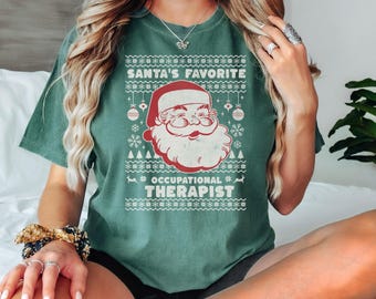 May include: Light green t-shirt with a festive design. The graphic features Santa Claus with the text "SANTA'S FAVORITE OCCUPATIONAL THERAPIST" in a Christmas sweater style. The shirt is a solid color and has a relaxed fit.