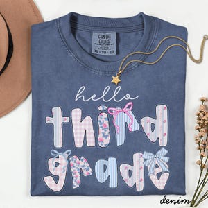 Könnte beinhalten: Ein denimblaues Comfort Colors T-Shirt mit dem Schriftzug "hello third grade" in einer verspielten Schriftart. Die Buchstaben sind mit floralen, Vichy- und Streifenmustern verziert. Eine goldene Sternkette und ein brauner Hut sind ebenfalls zu sehen.