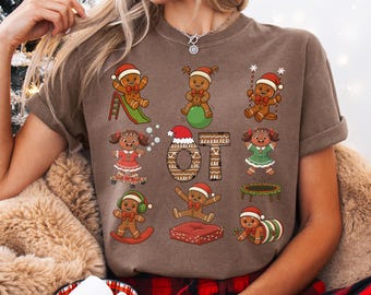 May include: Brown t-shirt with a gingerbread theme. The shirt features gingerbread people in various poses, including on a slide, balancing on a ball, and on a trampoline. The text "OT" is also displayed on the shirt.