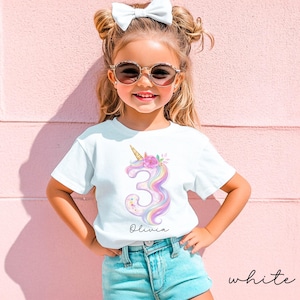 May include: A white t-shirt featuring a colorful unicorn design with the number 3 and the name "Olivia". The unicorn has a golden horn and is decorated with flowers. The shirt is worn by a young girl wearing sunglasses and a white bow.