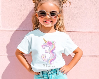 May include: A white t-shirt featuring a colorful unicorn design with the number 3 and the name "Olivia". The unicorn has a golden horn and is decorated with flowers. The shirt is worn by a young girl wearing sunglasses and a white bow.