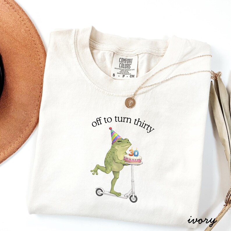May include: Ivory-colored t-shirt with the phrase "off to turn thirty" printed on the front. The graphic features a green frog wearing a party hat and riding a scooter while holding a birthday cake with the number 30. A gold necklace is draped on the shirt.
