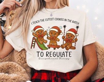 May include: Ivory-colored t-shirt featuring a playful graphic of three gingerbread men wearing Santa hats. The text reads "I teach the cutest cookies in the batch" and "To Regulate Occupational Therapy". The gingerbread men are on a slide, skateboard, and swing.