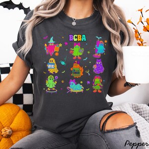 Könnte beinhalten: Dunkelgraues T-Shirt mit einer farbenfrohen Grafik von Cartoon-Geistern und dem Text "BCBA" darüber. Die Geister sind in verschiedenen Posen dargestellt, einige halten Gegenstände wie Bücher und Blumen. Das T-Shirt hat einen lässigen Vintage-Look.