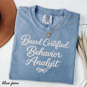 Könnte beinhalten: Ein blaues Comfort Colors T-Shirt mit den Worten "Board Certified Behavior Analyst" in Weiß gestickt. Eine goldene Halskette mit Anhänger liegt über dem Shirt. Das Shirt ist auf einer weißen Oberfläche gefaltet.