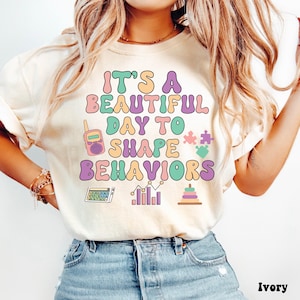 Könnte beinhalten: Elfenbeinfarbenes T-Shirt mit dem Text "It's a beautiful day to shape behaviors" in Pastellfarben. Das Design umfasst ein Walkie-Talkie, Puzzleteile, einen Taschenrechner, ein Diagramm und einen Ringturm.