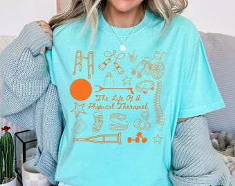 Puede incluir: Una camiseta turquesa con el texto "The Life Of A Physical Therapist" e ilustraciones naranjas de herramientas de fisioterapia. El diseño incluye una silla de ruedas, pesas y una columna vertebral.