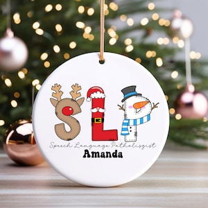 May include: A white ceramic Christmas ornament with the letters "SLP" decorated as a reindeer, Santa, and snowman. The text "Speech Language Pathologist" and a name are below the letters. A gold string is attached.