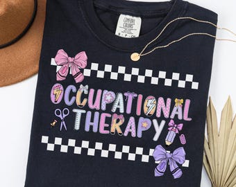May include: Black t-shirt with the words "OCCUPATIONAL THERAPY" in colorful letters and graphics. The design includes bows, scissors, and a pencil. A gold necklace and a brown hat are also visible.