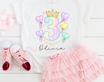 May include: White t-shirt with a colorful number 3 design, a gold crown, and heart-shaped balloons. The name "Olivia" is written in script below the design. A pink tulle skirt and pink sneakers complete the outfit.
