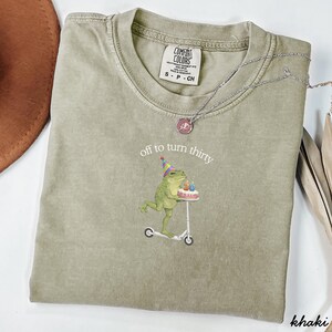 May include: Khaki-colored t-shirt with the text "off to turn thirty" and a graphic of a frog on a scooter holding a birthday cake. The shirt has a crew neck and short sleeves. A silver necklace is draped across the shirt.