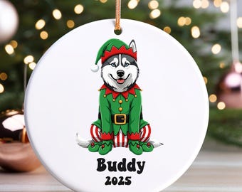 May include: A white ceramic Christmas ornament featuring a Siberian Husky dressed as an elf. The dog wears a green outfit with red and white striped socks and a matching hat. The text "Buddy 2025" is printed below the dog.