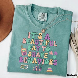 Op de afbeelding: Zeegroen T-shirt met de tekst "It's a beautiful day to shape behaviors" in kleurrijke letters. Het shirt heeft verschillende illustraties, waaronder een rekenmachine, een grafiek, een walkie-talkie, een puzzelstukje en een stapelbaar speelgoed.