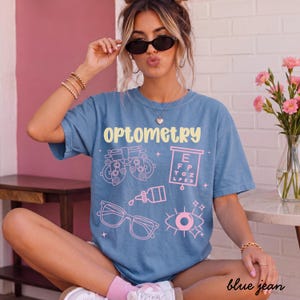 May include: A blue short-sleeve t-shirt featuring the word OPTOMETRY in yellow text. The shirt includes light pink graphic illustrations of an eye exam phoropter, a Snellen eye chart, a bottle of eye drops, eyeglasses, and an eye icon.