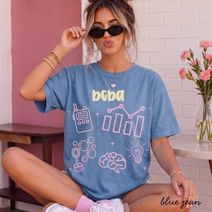 Könnte beinhalten: Ein blaues Kurzarm-T-Shirt mit dem gelben Schriftzug "bcba" und verschiedenen rosa Grafiksymbolen, darunter ein Funkgerät, ein Balkendiagramm, ein Seifenblasenstab, ein menschliches Gehirn und eine Glühbirne auf der Vorderseite.