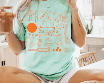 Puede incluir: Camiseta de color verde azulado claro con ilustraciones naranjas de herramientas de fisioterapia y el texto "The Life Of A Physical Therapist". La camiseta tiene cuello redondo y mangas cortas. La persona está sentada y sosteniendo un vaso de café helado.