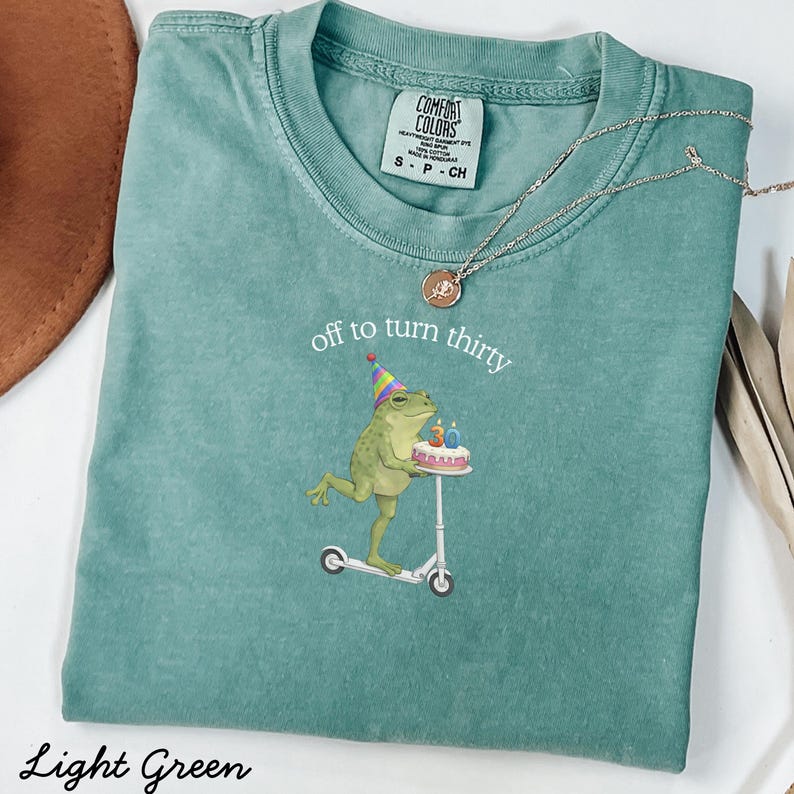 May include: Light green t-shirt with the text "off to turn thirty" and a graphic of a frog riding a scooter while holding a birthday cake. The shirt has a crew neck and short sleeves. A silver necklace is draped across the shirt.