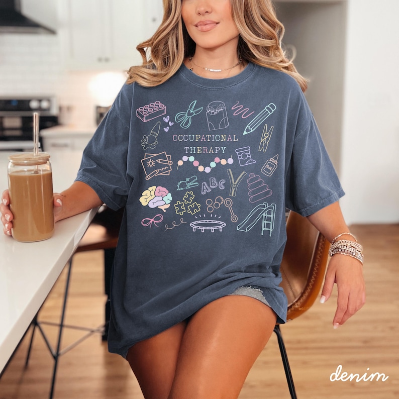 Pu&ograve; includere: Una maglietta blu denim con le parole "OCCUPATIONAL THERAPY" e illustrazioni colorate di strumenti terapeutici. La maglietta ha un girocollo e maniche corte. La parola "denim" &egrave; stampata in basso a destra.