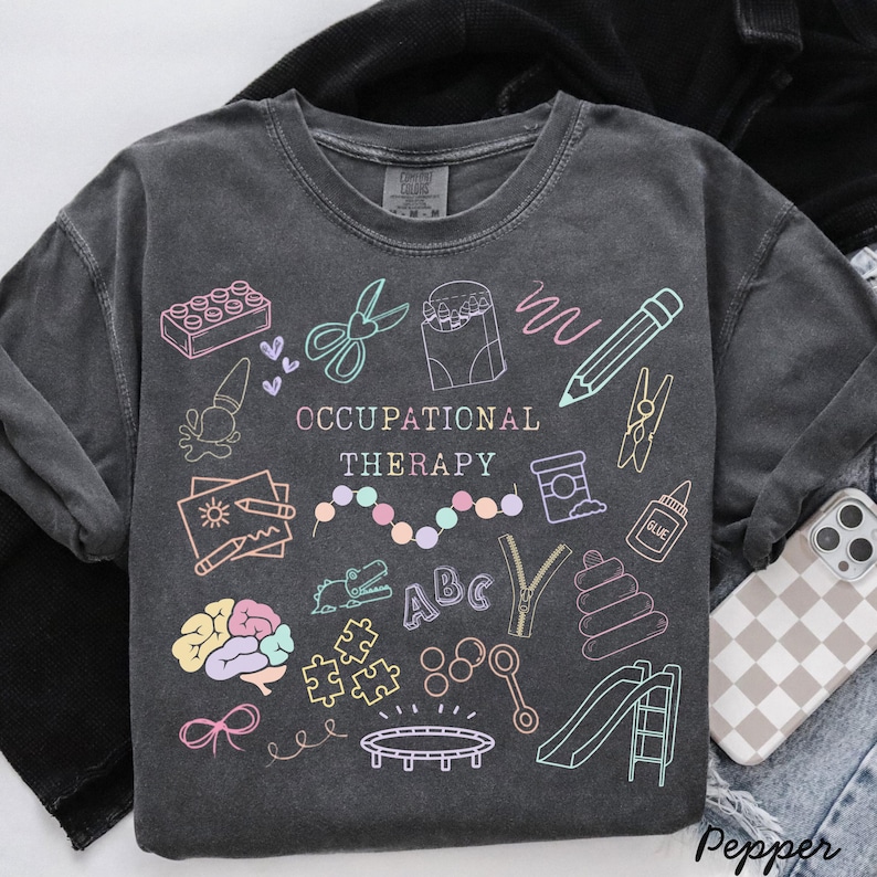 Pu&ograve; includere: Felpa grigio scuro con la scritta "OCCUPATIONAL THERAPY" e illustrazioni colorate di strumenti terapeutici. Il design include forbici, una matita, un cervello e uno scivolo. La felpa &egrave; uno stile casual e confortevole.