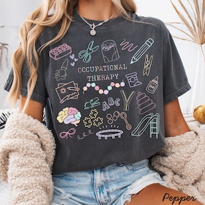 Pu&ograve; includere: T-shirt grigio scuro con la scritta "OCCUPATIONAL THERAPY" e illustrazioni colorate di strumenti terapeutici. Il design include forbici, un cervello, pezzi di puzzle e uno scivolo.