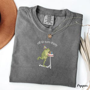 May include: A gray Comfort Colors t-shirt with the phrase "off to turn thirty" and a graphic of a frog riding a scooter while holding a birthday cake. The shirt has a crew neck and short sleeves. A silver necklace is draped across the shirt.
